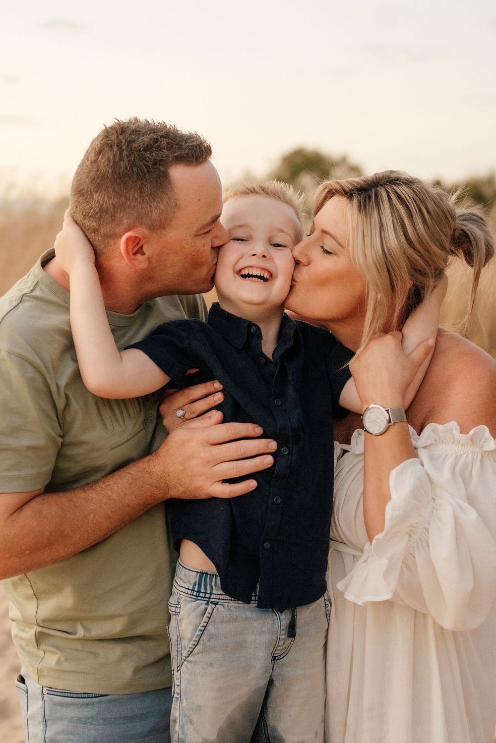 Parents kissing laughing young boy on cheeks in golden field at sunset.