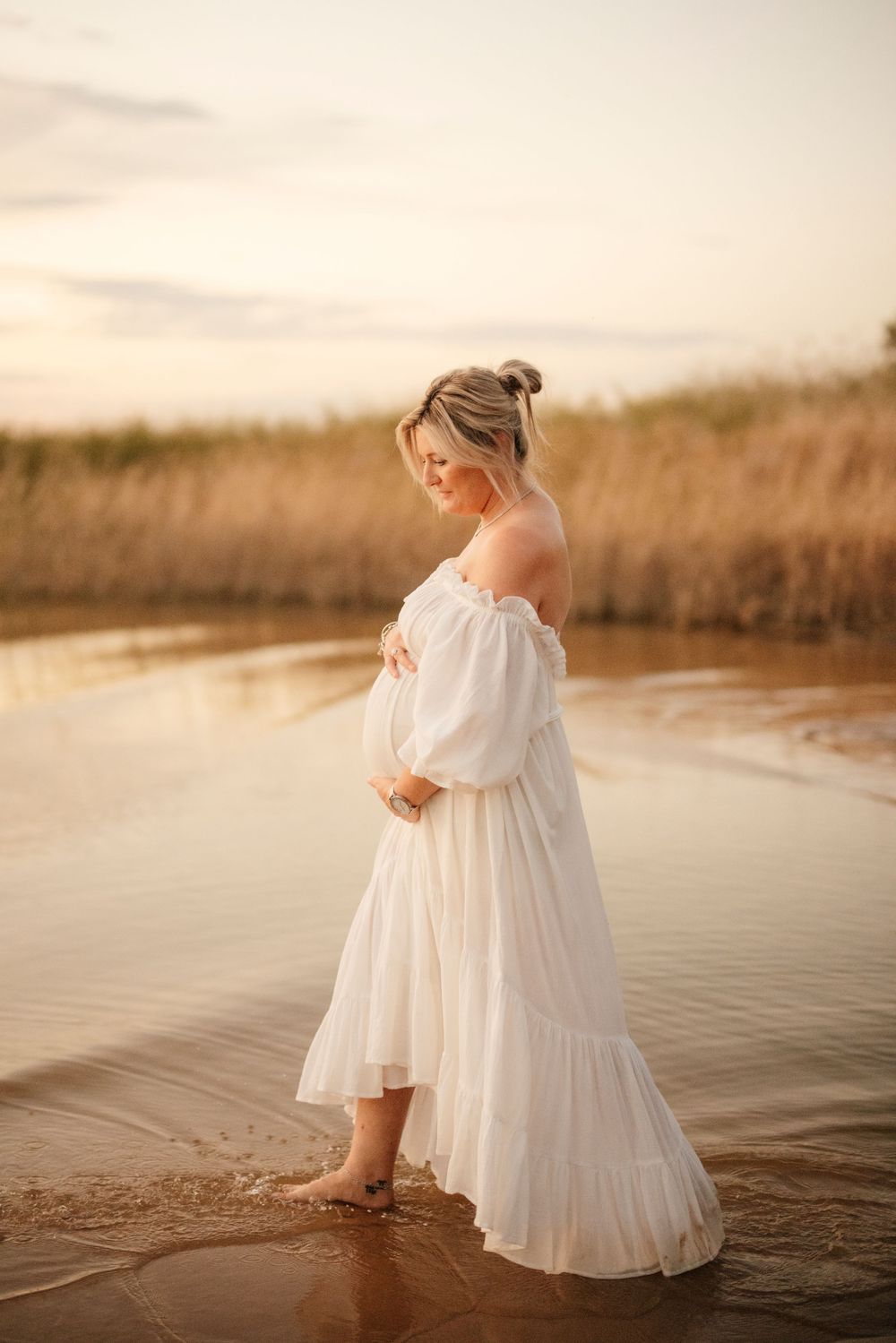 Pregnant woman in white off-shoulder dress standing barefoot in shallow water at sunset.