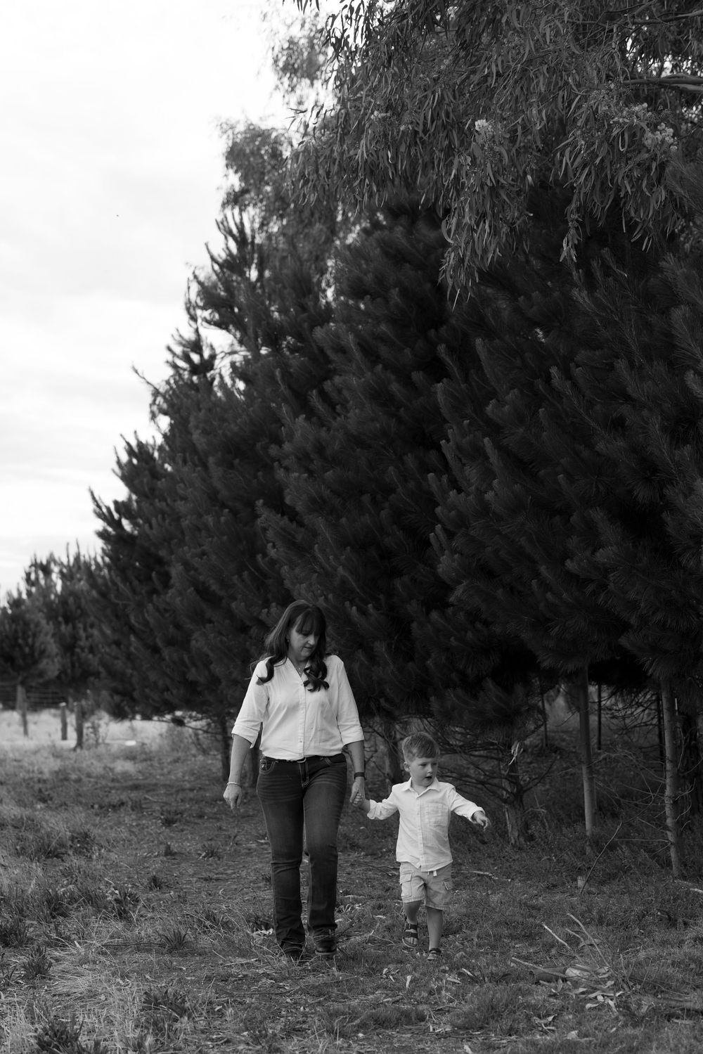 Black and white photo of mother and toddler walking hand in hand near trees.