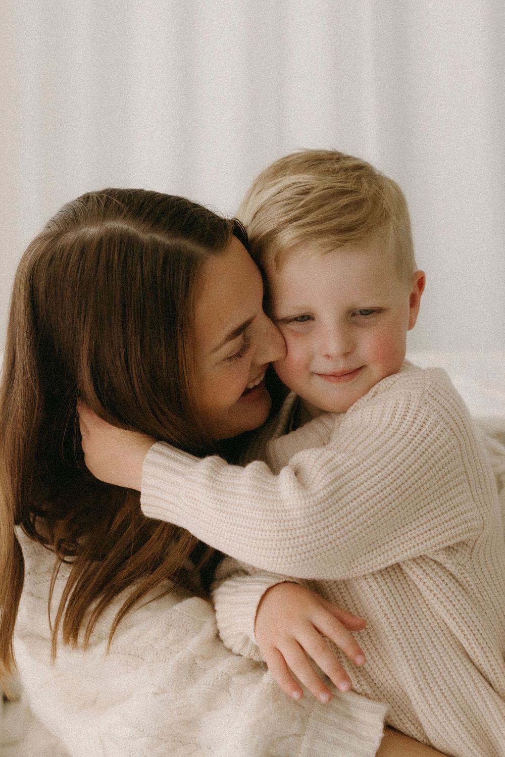 A dark-haired woman tenderly embraces a smiling blonde boy in matching cream knit sweaters.