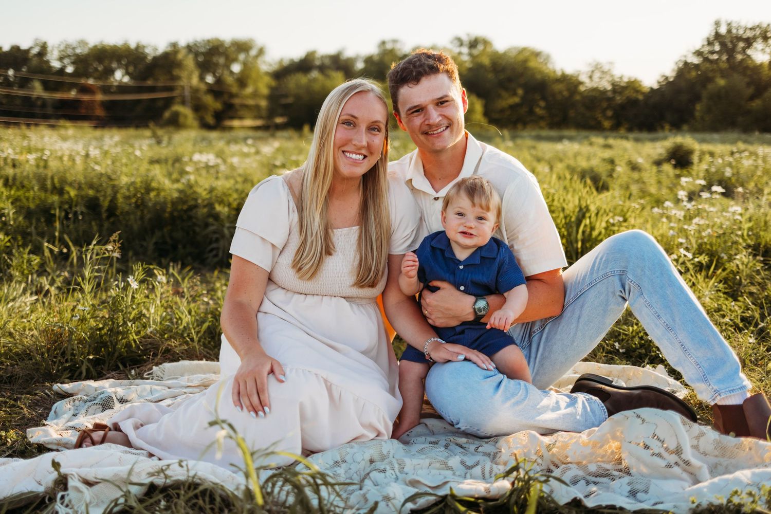 Happy family of three sitting on a blanket in a sunny green field, smiling couple with baby boy.