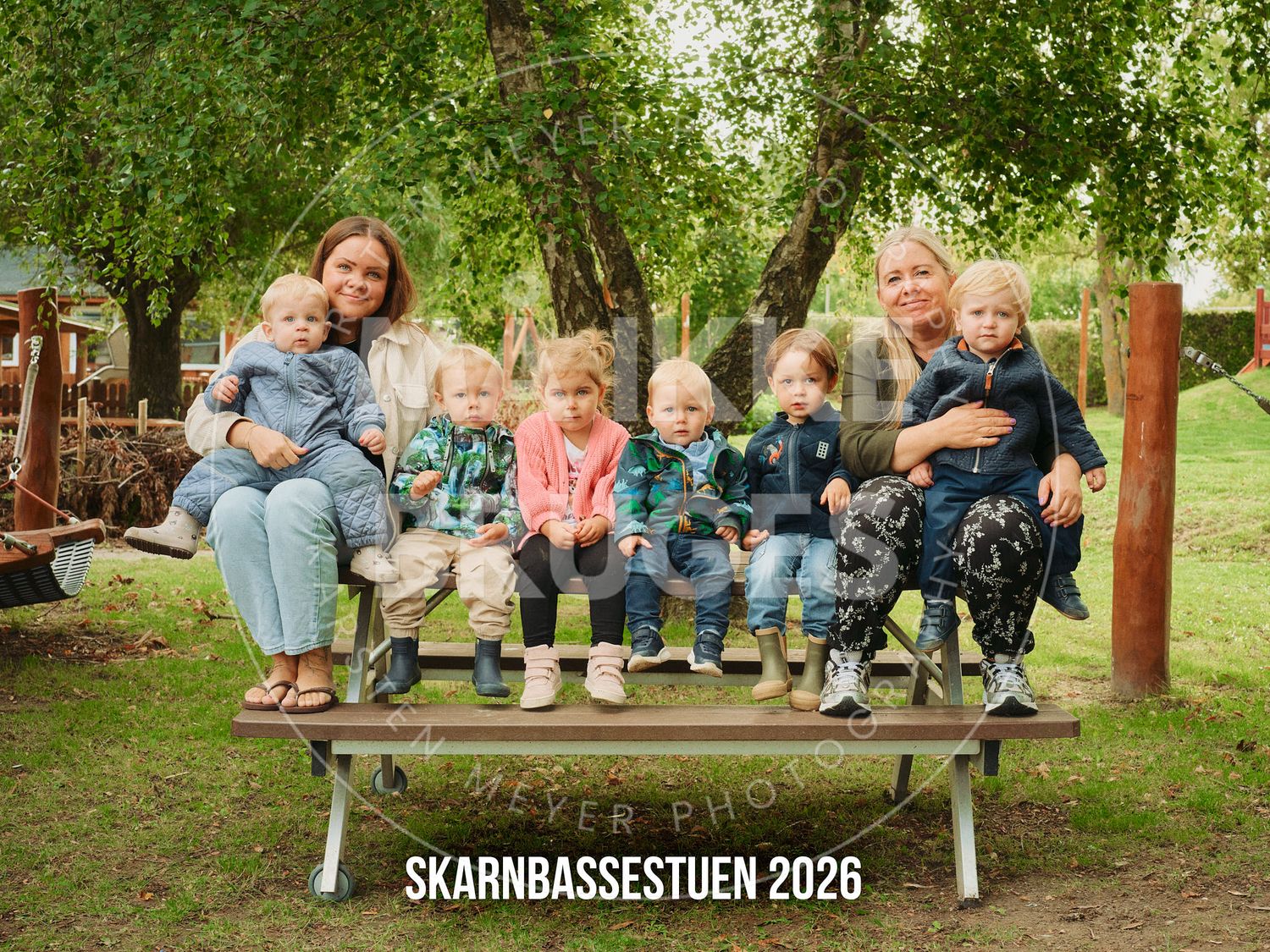 Two women and five young children sit together on a bench outdoors in a green garden setting, photo labeled Skarnbassestuen 2026.