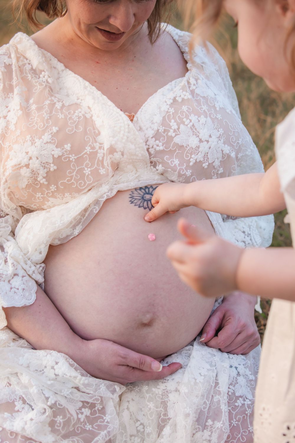 Pregnant woman in lace dress with child touching her baby bump, sunflower tattoo visible, outdoor maternity photo in Rouse Hill.