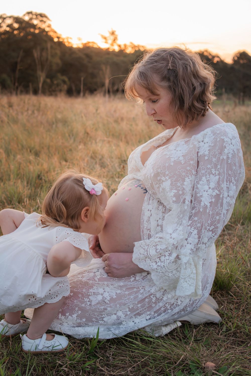 Pregnant woman in white lace dress sitting in golden field at sunset with toddler daughter kissing her baby bump in Rouse Hill.