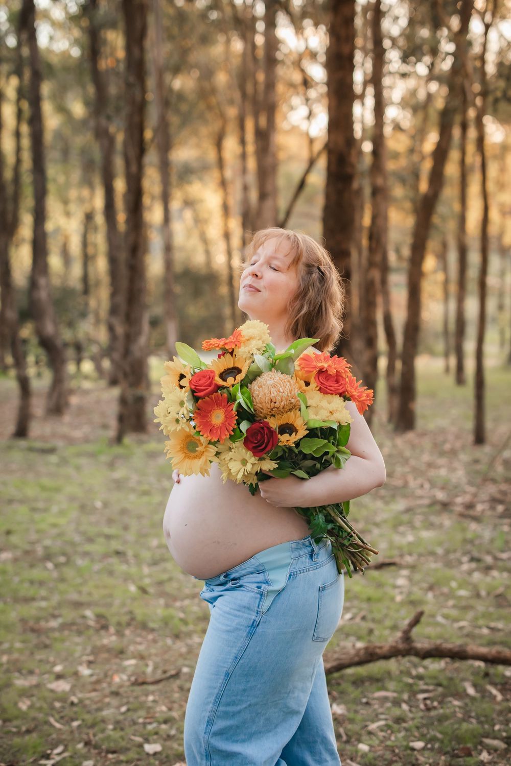 Pregnant woman in jeans holding colorful bouquet of orange and yellow flowers in a sunlit forest setting.
