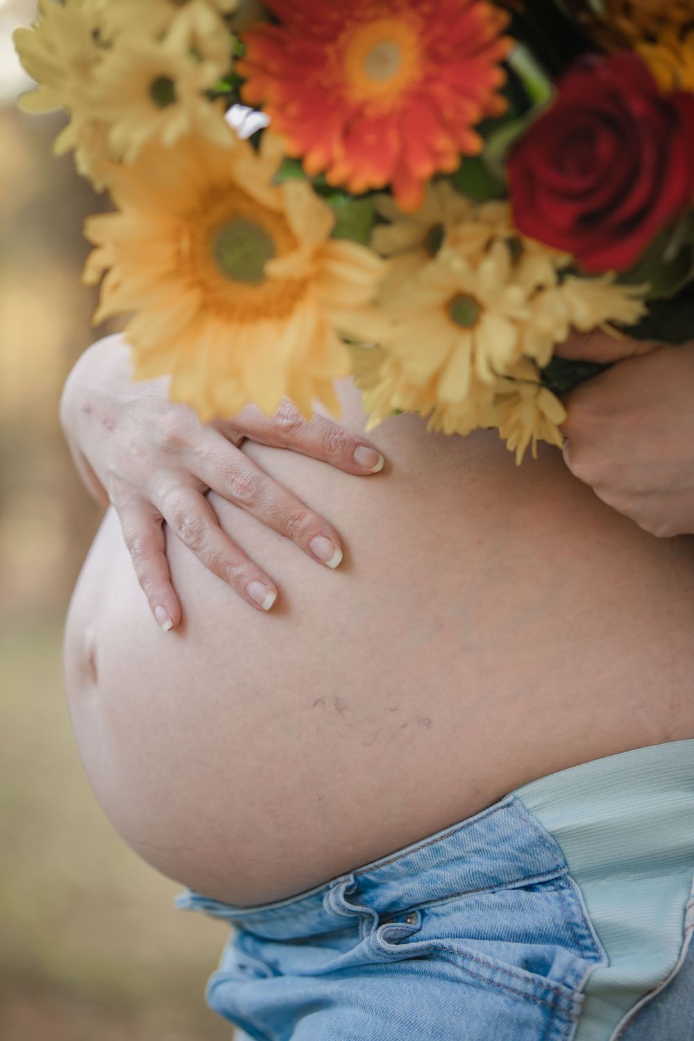 Pregnant woman cradling bare baby bump with hands, holding colorful bouquet of sunflowers and red roses outdoors.