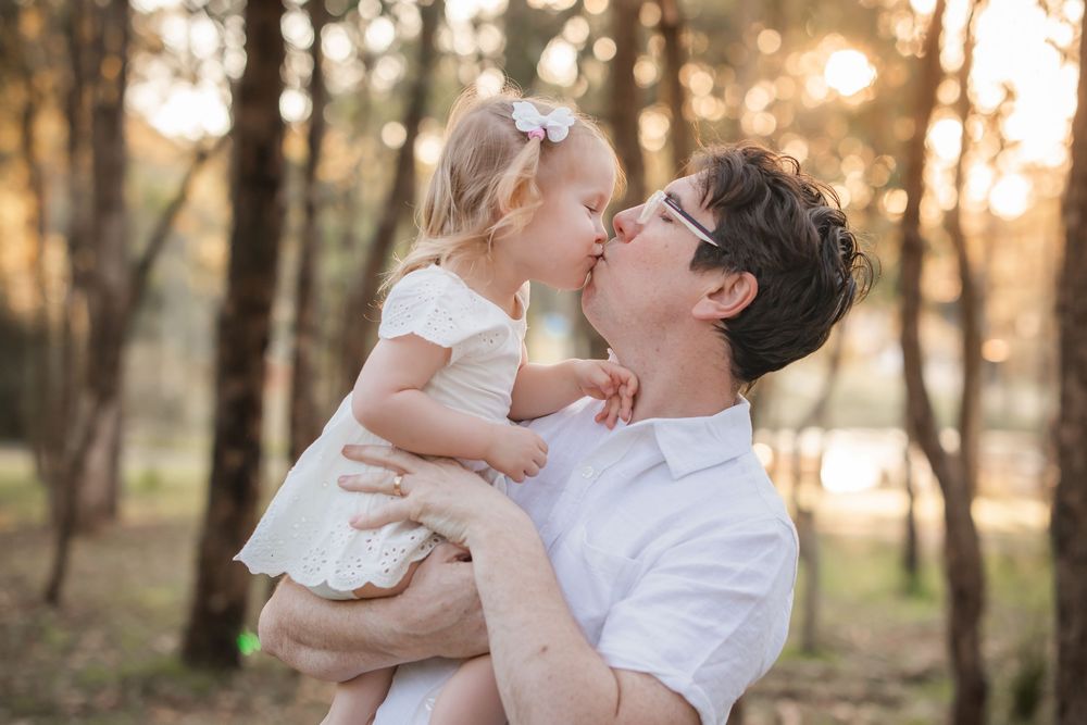 A father holds and kisses his young daughter in a sunlit forest, both dressed in white.