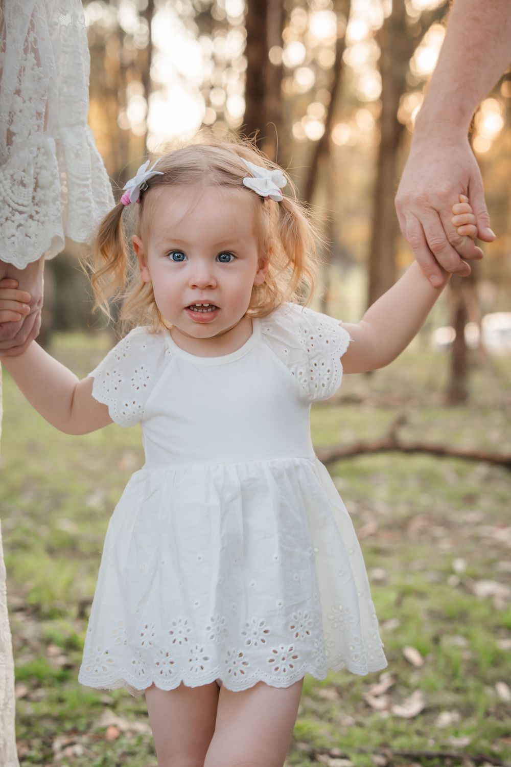 Toddler girl in white eyelet dress holds parents hands outdoors in sunlit forest setting.