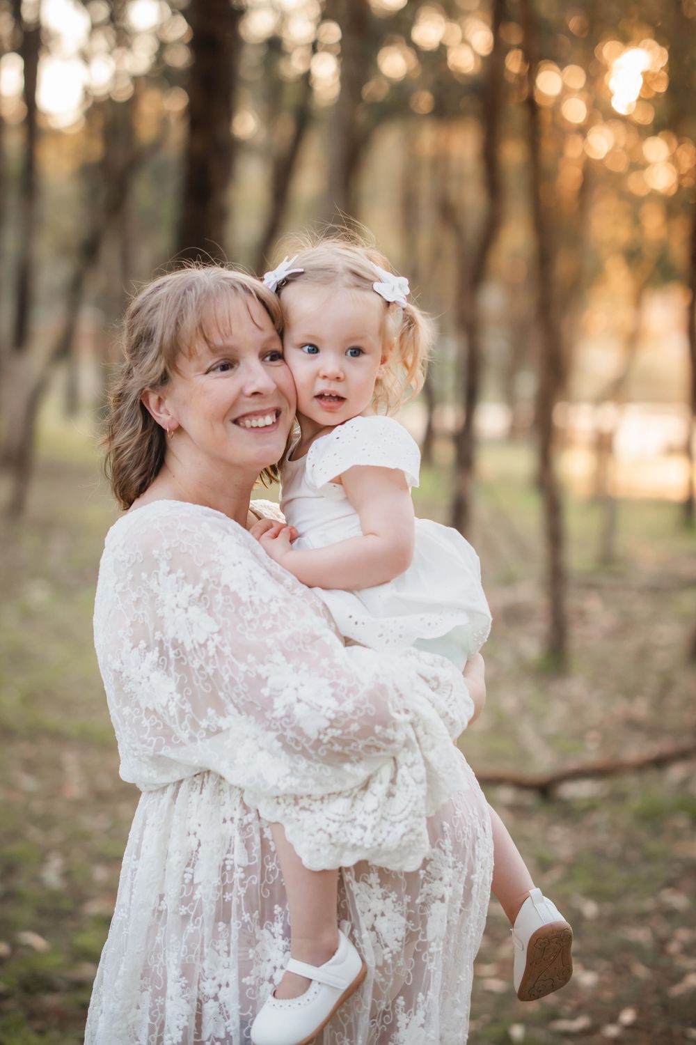A smiling woman in white lace dress holds a toddler girl in white dress outdoors in a sunlit forest setting.