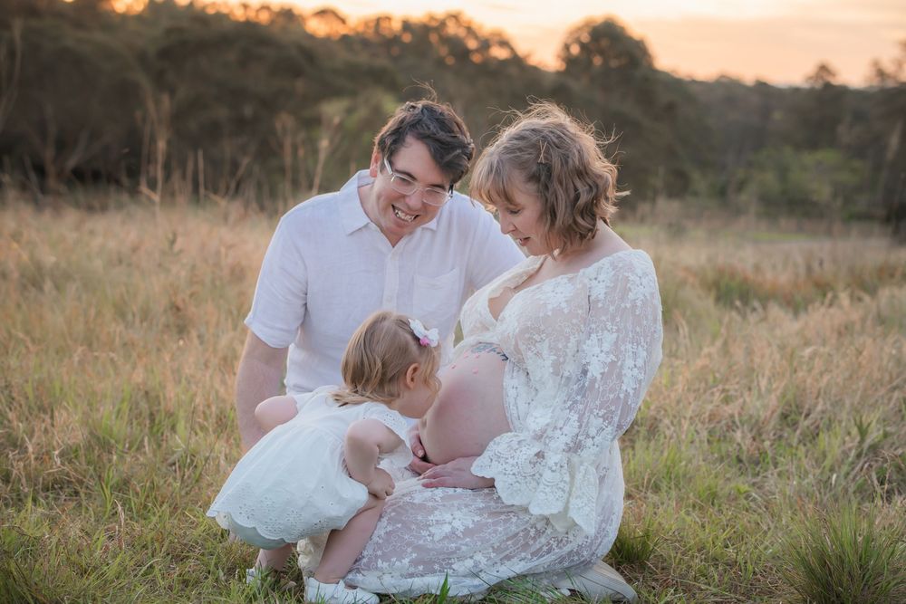 Pregnant woman in lace dress with husband and toddler daughter in golden field at sunset maternity photo.