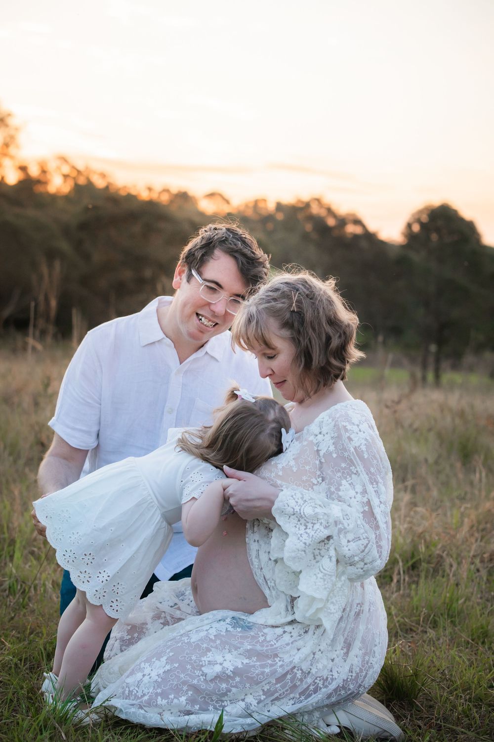 A smiling family shares a tender moment at sunset in a grassy field, with a young girl hugging her pregnant mother.