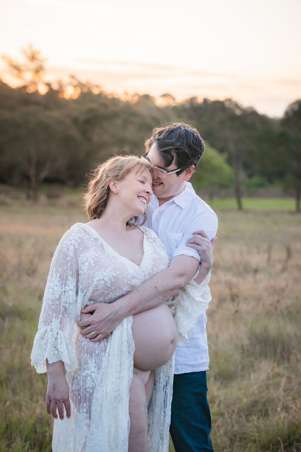 Couple sharing a tender maternity photography moment at golden hour at Rouse Hill Regional Park, woman in white lace gown showing baby bump.
