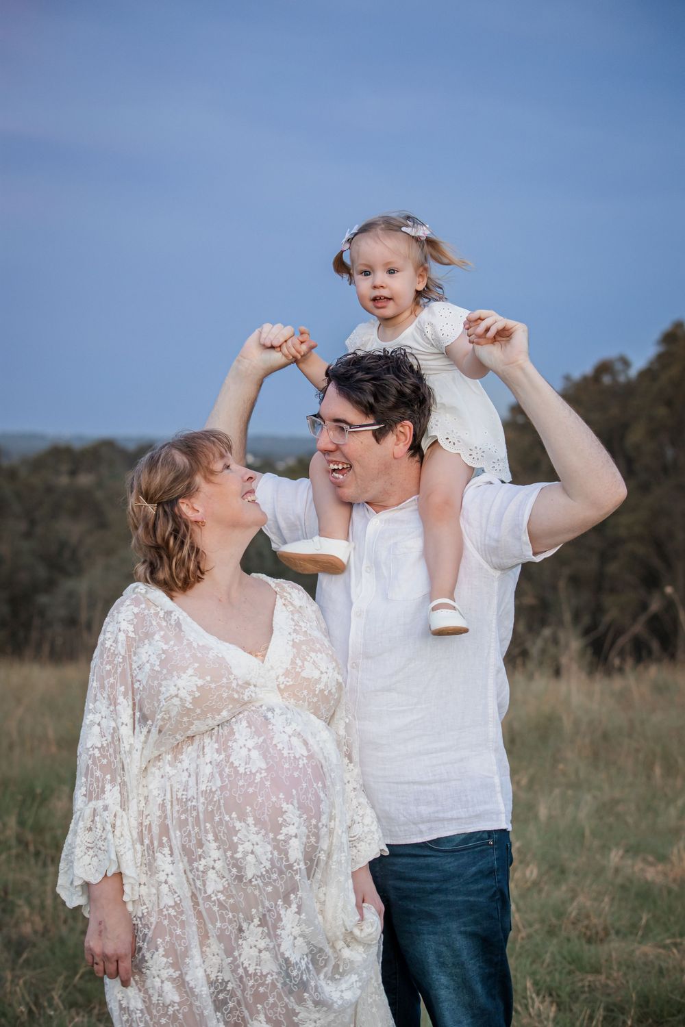 Pregnant woman in lace dress with husband and toddler daughter on shoulders in a sunny field at Rouse Hill Regional Park.