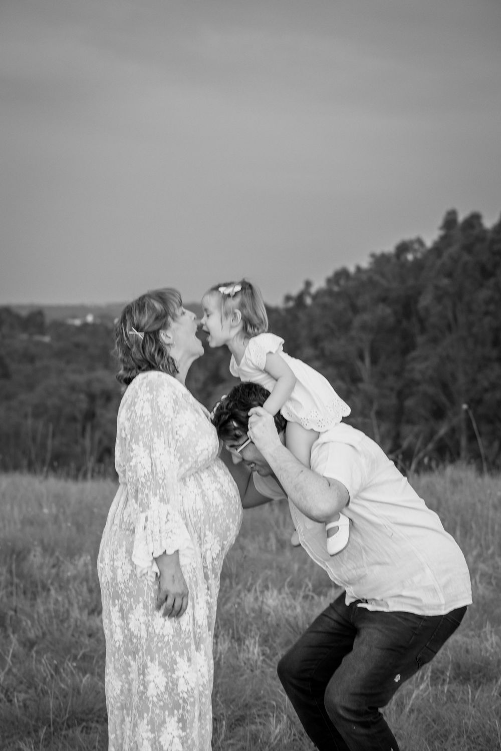 Black and white maternity photo of pregnant woman in lace dress with partner lifting toddler for a kiss outdoors.