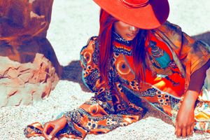 Bohemian woman in colorful printed dress and orange wide-brim hat sitting on sandy ground.
