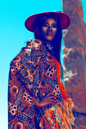 Stylish Black woman in vibrant tribal print cape and red wide-brim hat posing outdoors under bright blue sky.