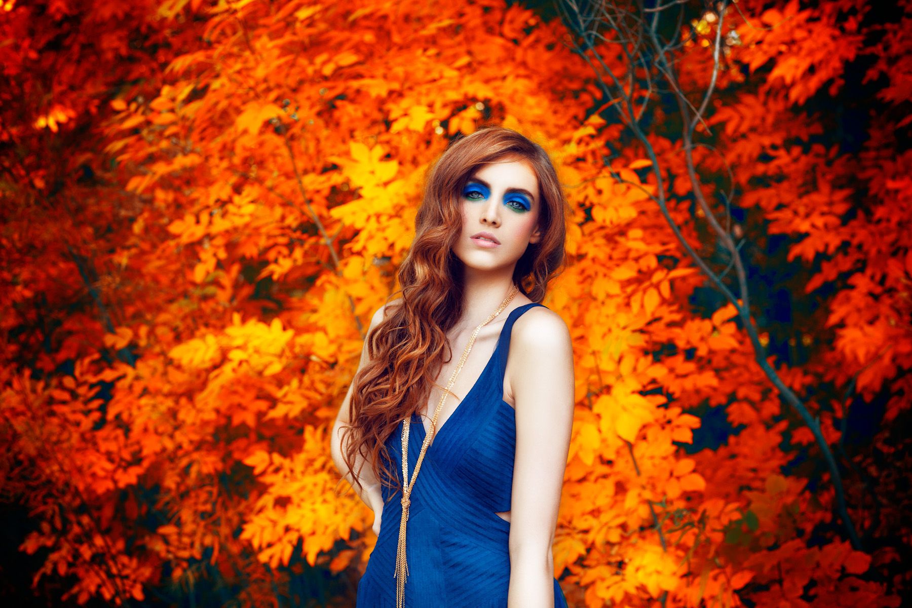 A woman with long red hair in a blue dress poses against vibrant orange autumn foliage.