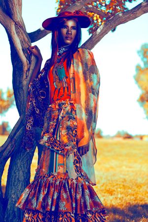 A model in colorful boho fashion with a wide-brim hat poses beside a tree in a golden field.