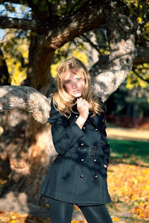 A blonde woman in a black coat stands between twisted tree branches in an autumn park.