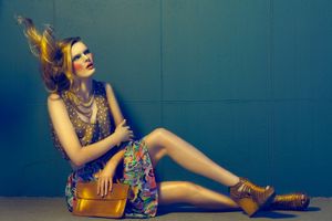 A fashion model in a colorful dress sits against a teal wall with a gold handbag.
