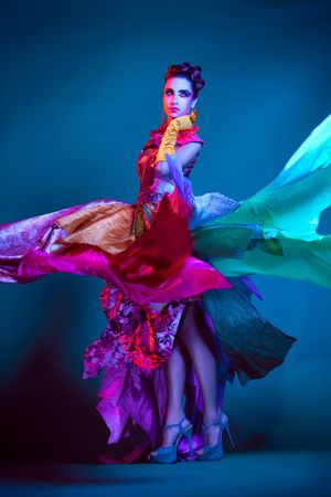 A woman in a vibrant flowing multi-colored gown dances under dramatic colorful studio lighting.