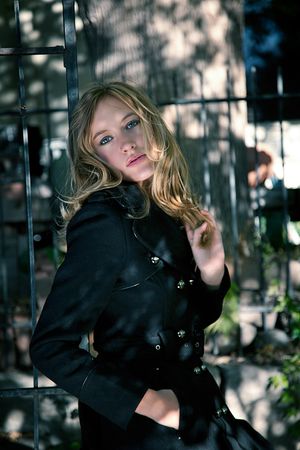 A blonde woman in a black coat leans against a metal gate in dappled sunlight.