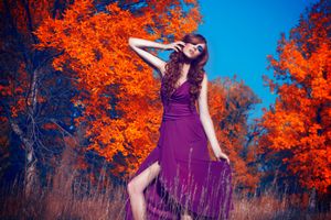 Woman in purple dress posing among vibrant orange autumn trees against blue sky.