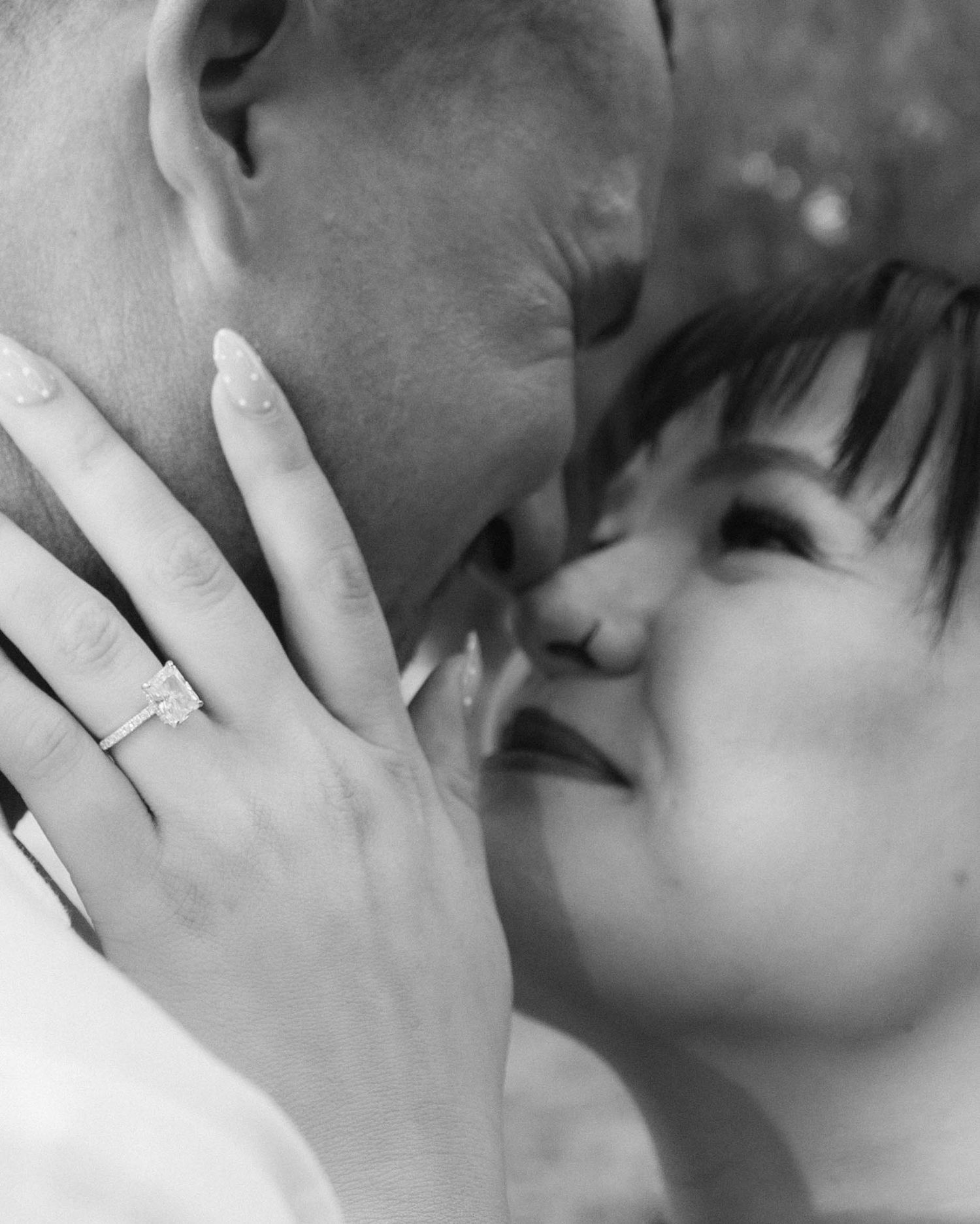 Black and white close-up of couple almost kissing, woman's hand showing elegant engagement ring with manicured nails.