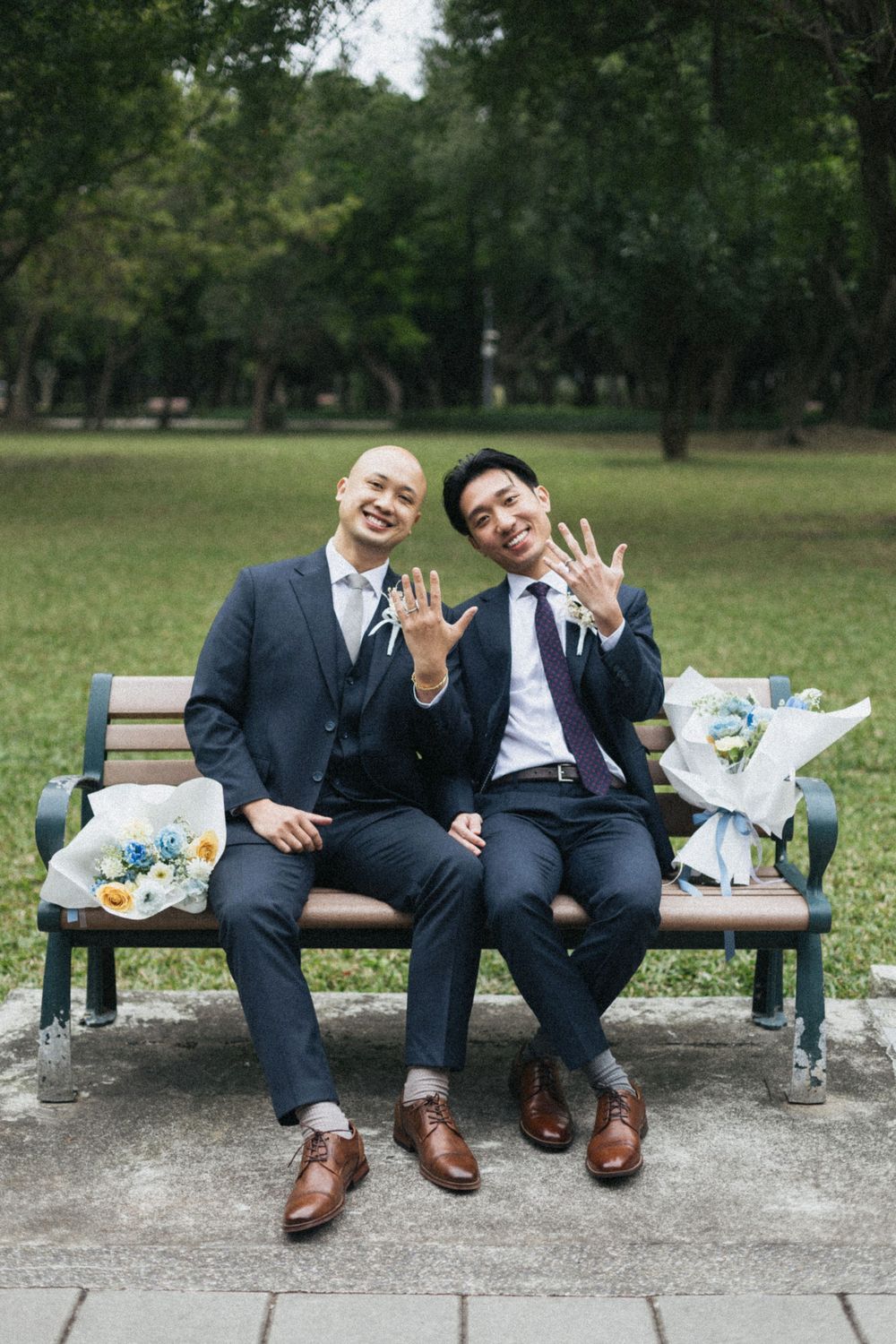 Two grooms in navy suits show off wedding rings on a park bench with floral bouquets beside them.