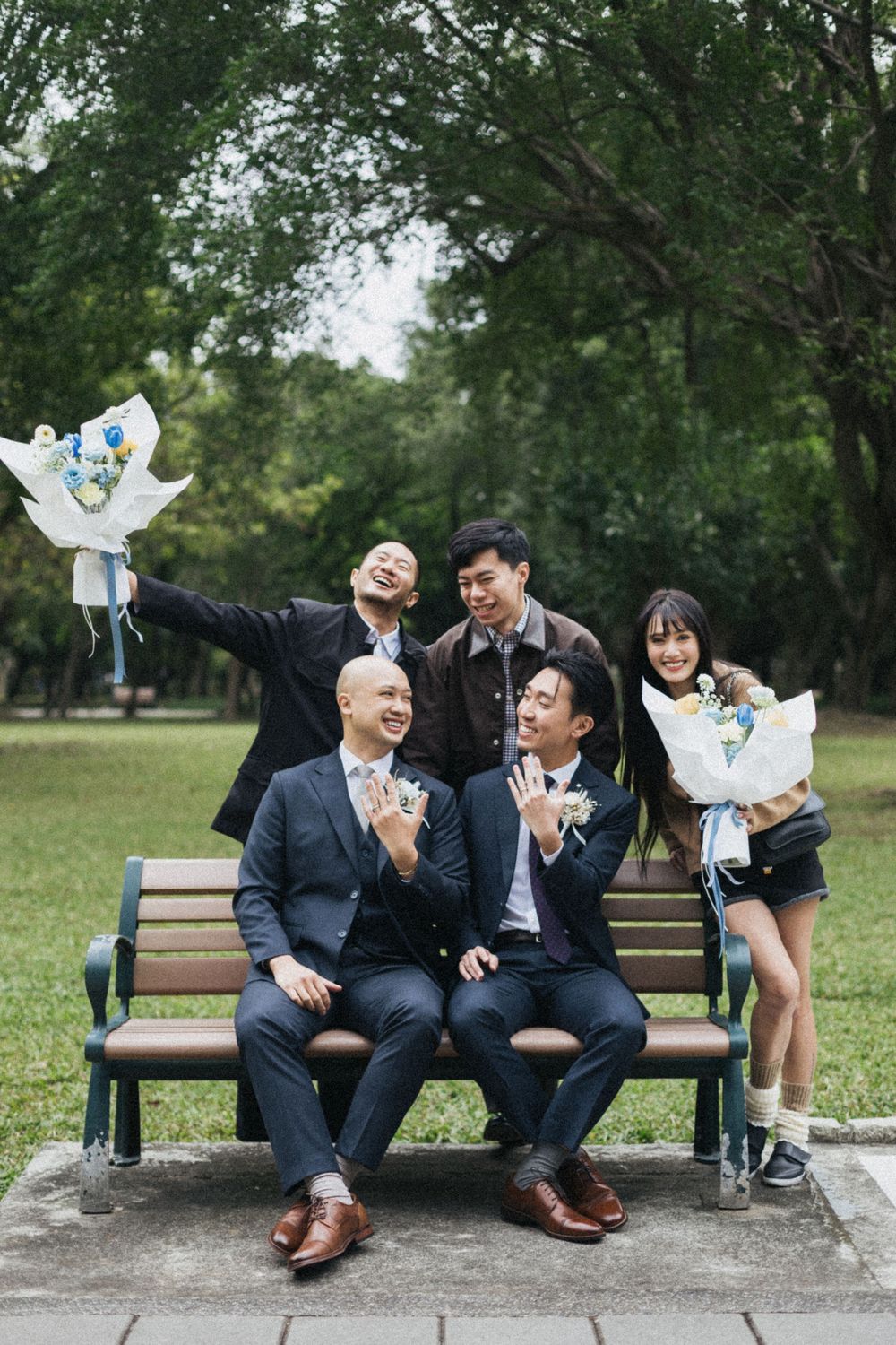 Five young Asian friends celebrate outdoors on a park bench, holding flower bouquets and showing rings in formal attire.