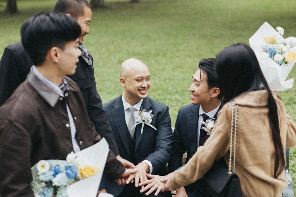 Groom in navy suit with boutonniere smiles with friends holding flower bouquets outdoors at a wedding celebration.