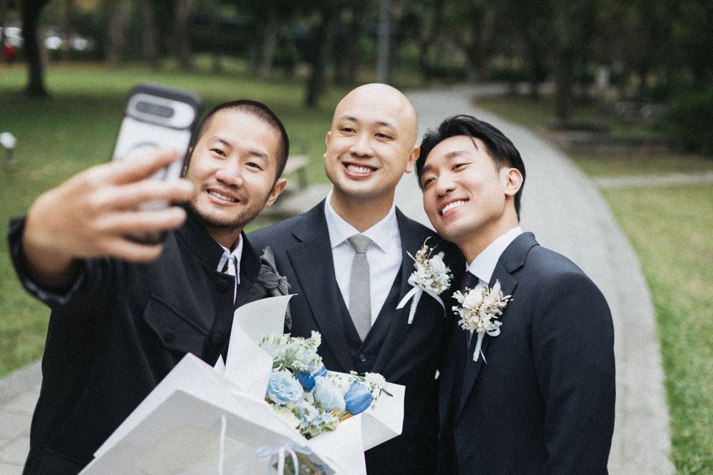Three Asian men in formal suits take a selfie outdoors, with one holding a bouquet of blue and white flowers.