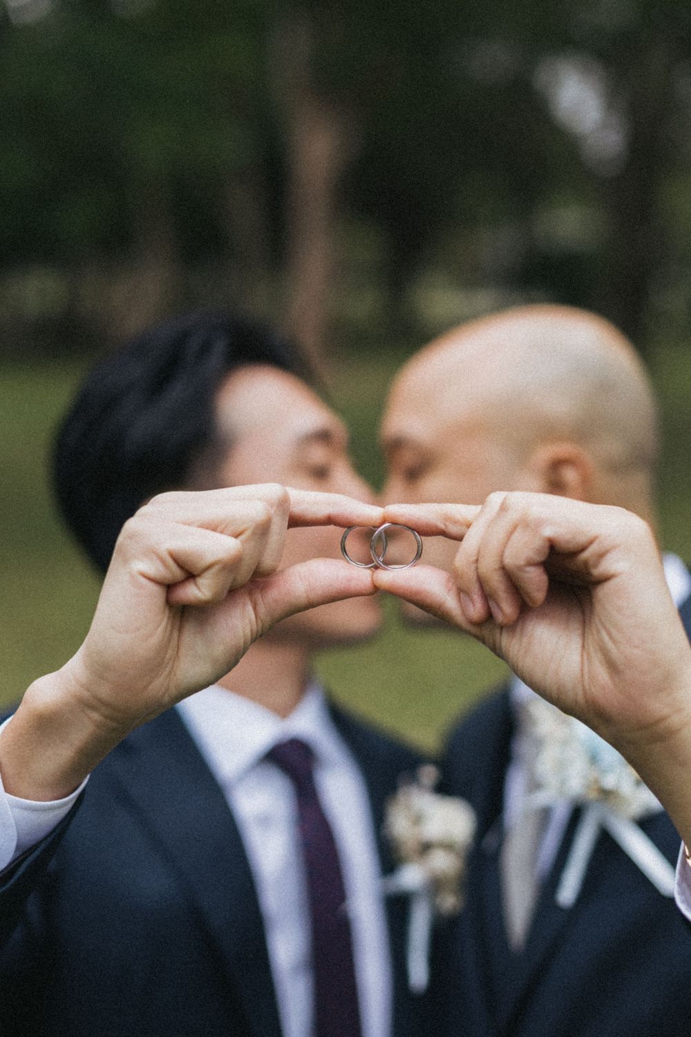 Two grooms in suits kiss while holding up their wedding rings together outdoors at their wedding ceremony.
