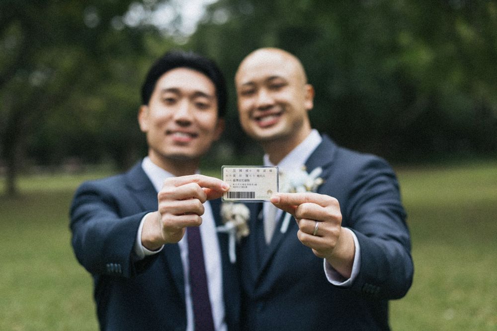 Two grooms in navy suits smiling and holding up their marriage license outdoors at their wedding ceremony.