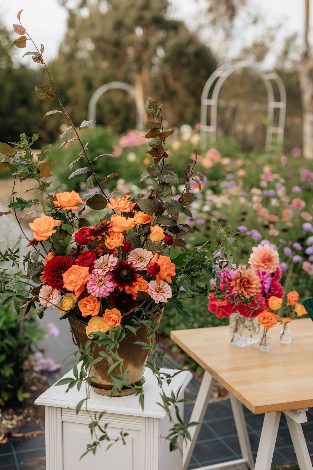 Lush floral arrangement with orange roses and red dahlias in a brass vase, set in a blooming garden.