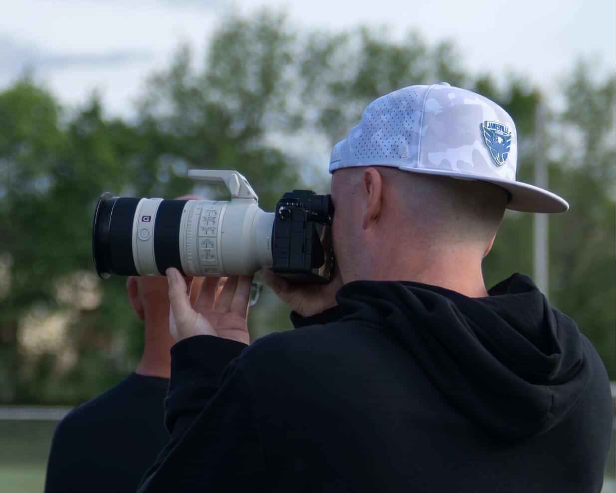 A photographer seen from behind in a white cap and black hoodie aims a Sony camera with a long white lens across a grassy field