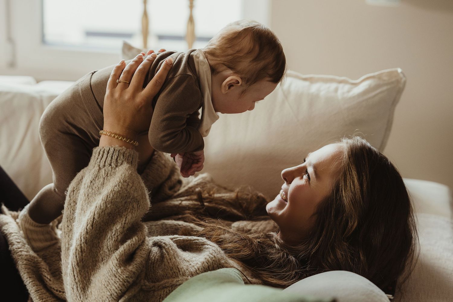 Mother lying on couch lifting smiling baby in the air during playful bonding moment at home.