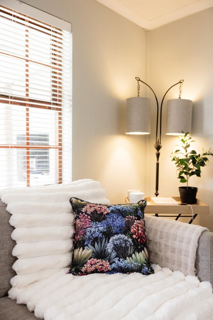 Cozy living room corner with floral pillow on white sofa, floor lamp, and wooden blinds.