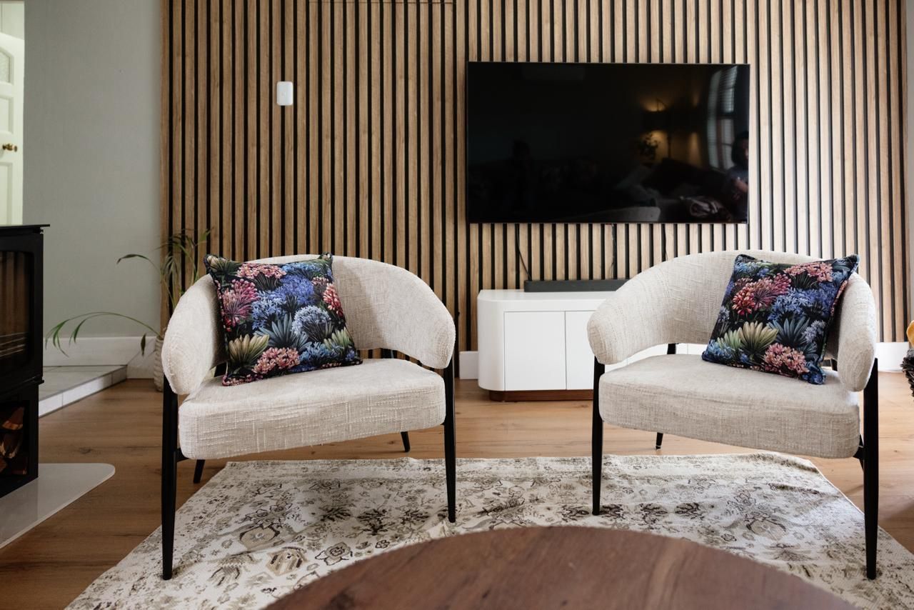 Two modern white boucle armchairs with floral cushions on a rug, set against a timber slat wall with mounted TV.