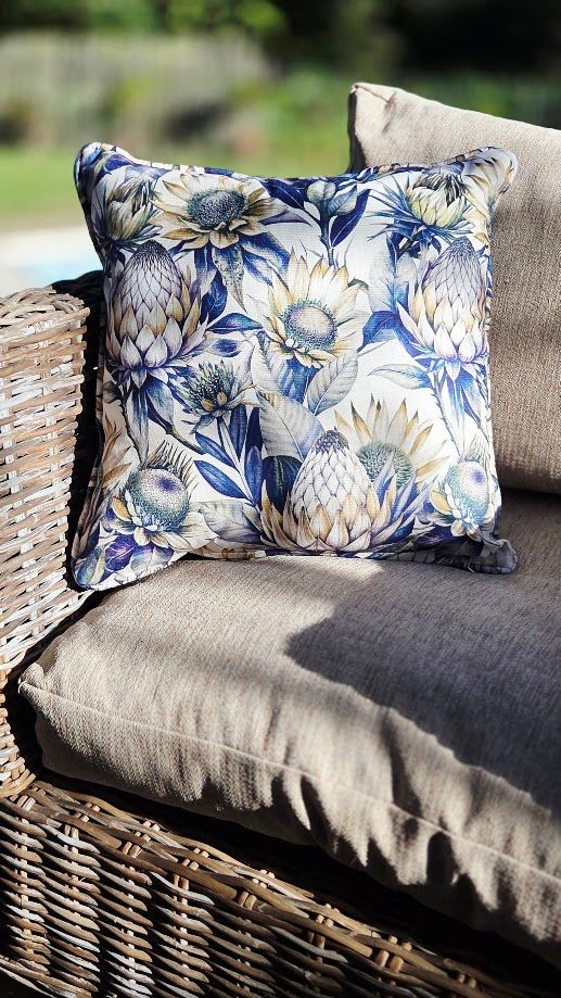 Floral protea print cushion in blue and cream tones on a wicker outdoor sofa with grey cushions.