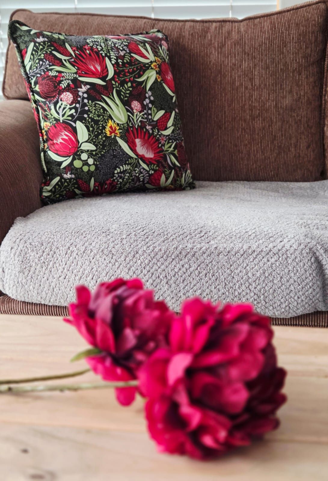 Cozy brown sofa with floral cushion and pink peonies on wooden table in stylish living room.