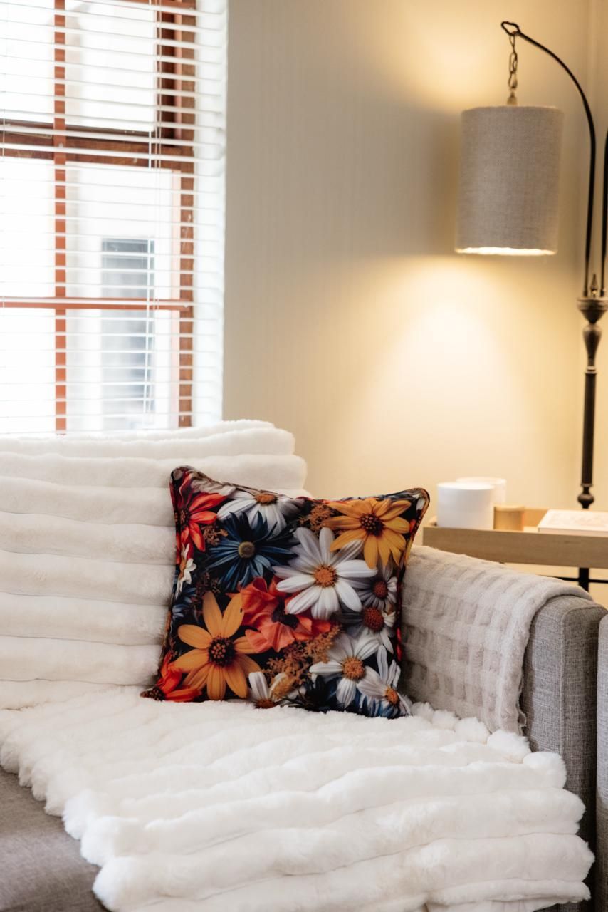 Cozy living room corner with a floral throw pillow on a white fluffy sofa near a window with blinds and a floor lamp.