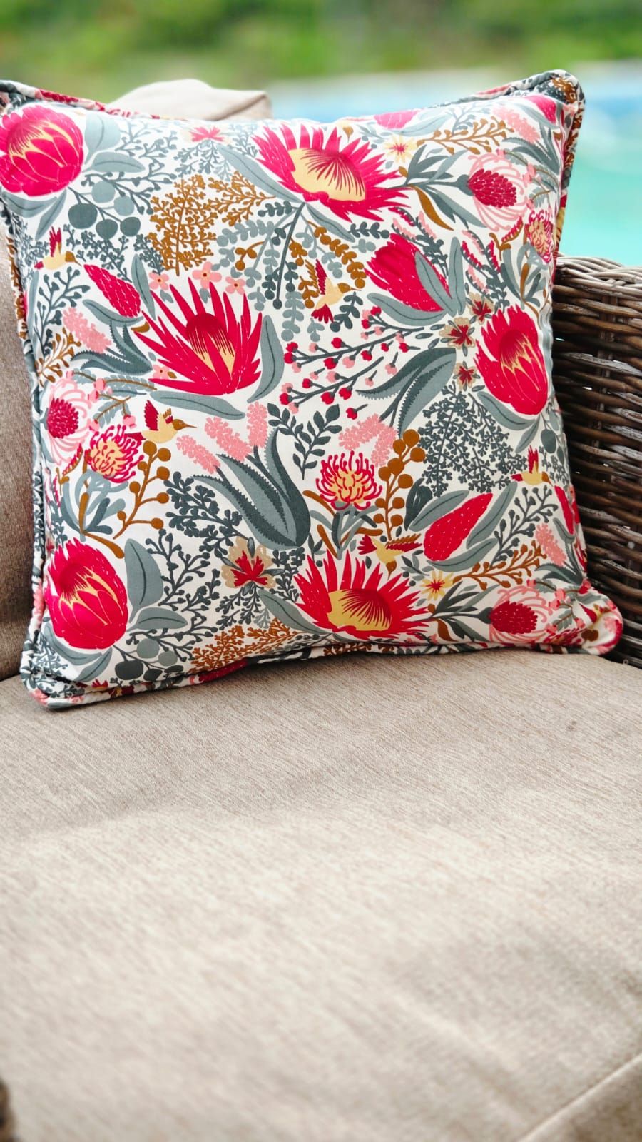 Vibrant floral throw pillow with pink protea blooms and grey foliage on a wicker outdoor chair.