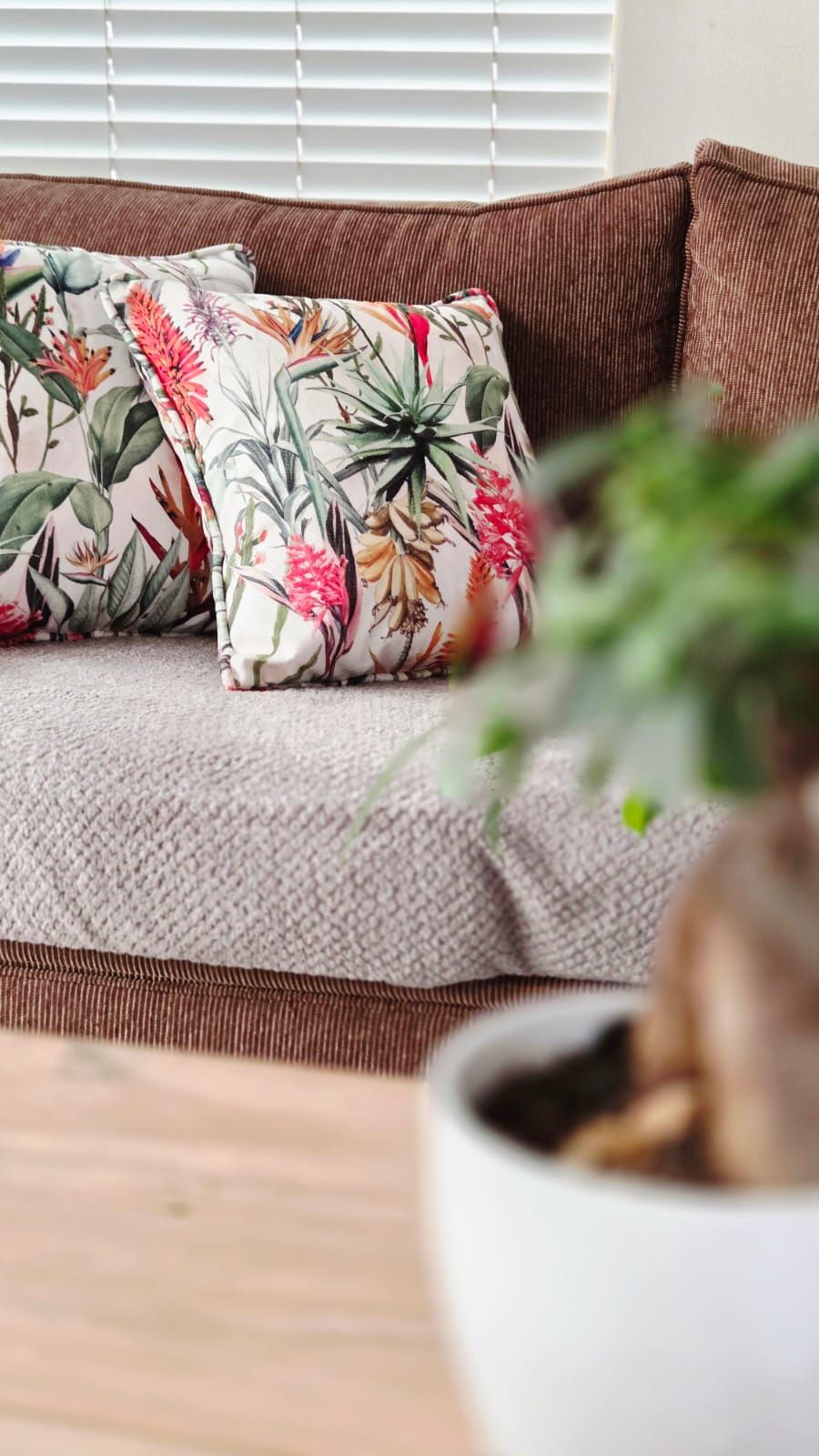 Brown sofa with floral tropical print cushions and grey throw, blurred green plant in white pot foreground.