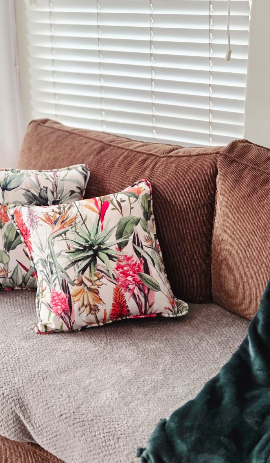 Brown sofa with two tropical floral throw pillows near a window with white blinds.