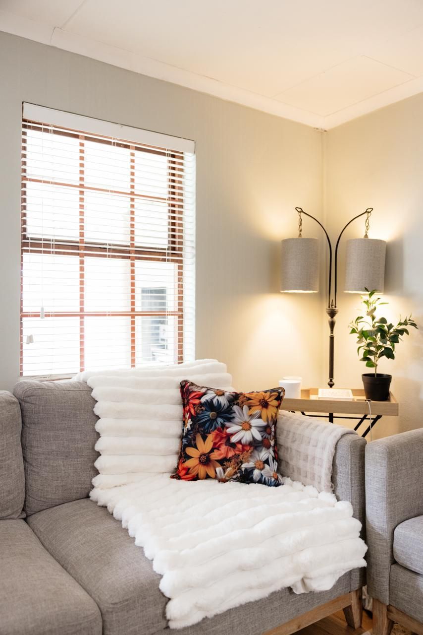 Cozy living room corner with gray sofa, white faux fur throw, floral pillow, floor lamp, and wooden blinds.