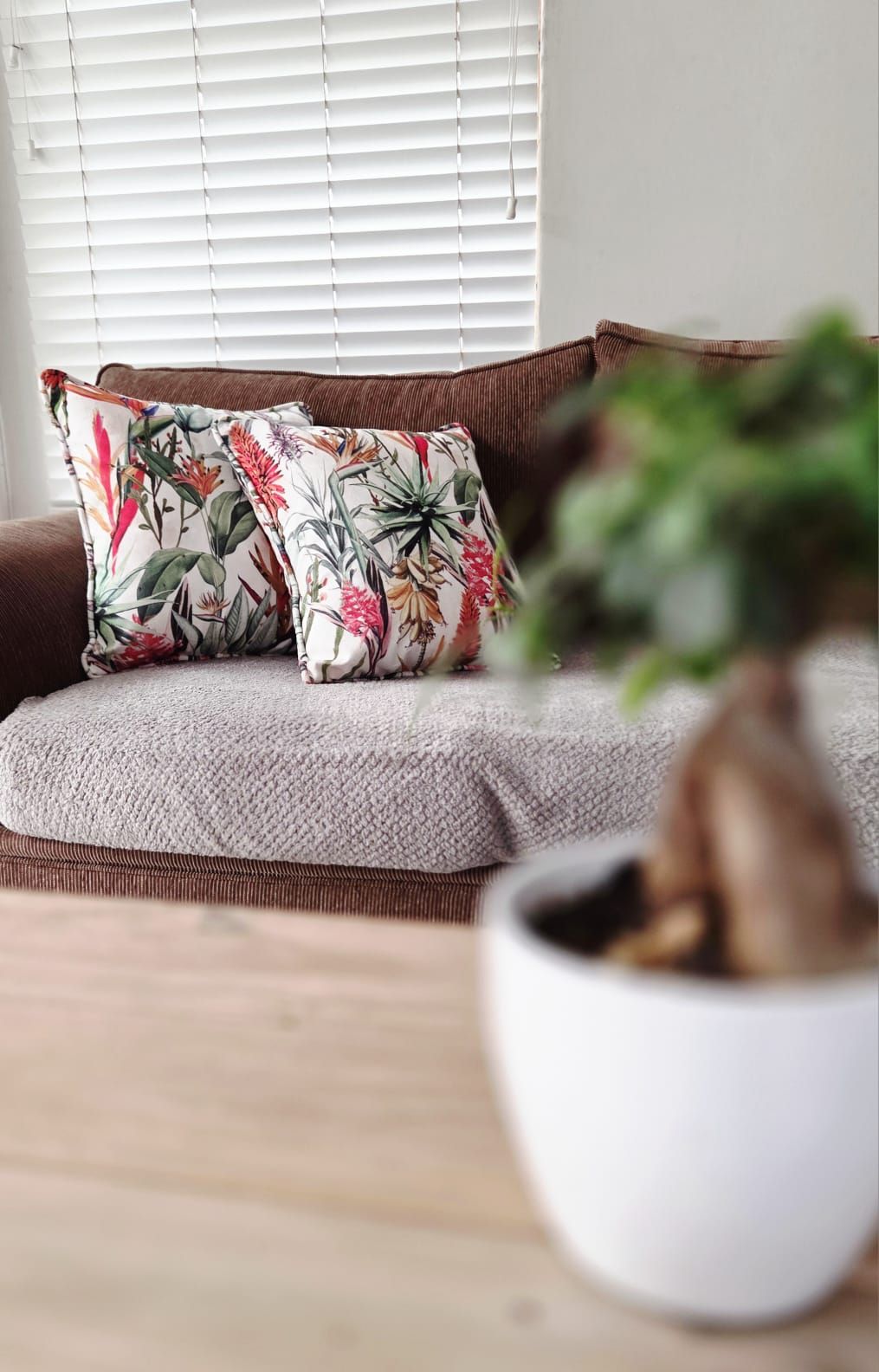 Cozy brown sofa with tropical floral cushion near window blinds, blurred green plant in foreground on wooden floor.