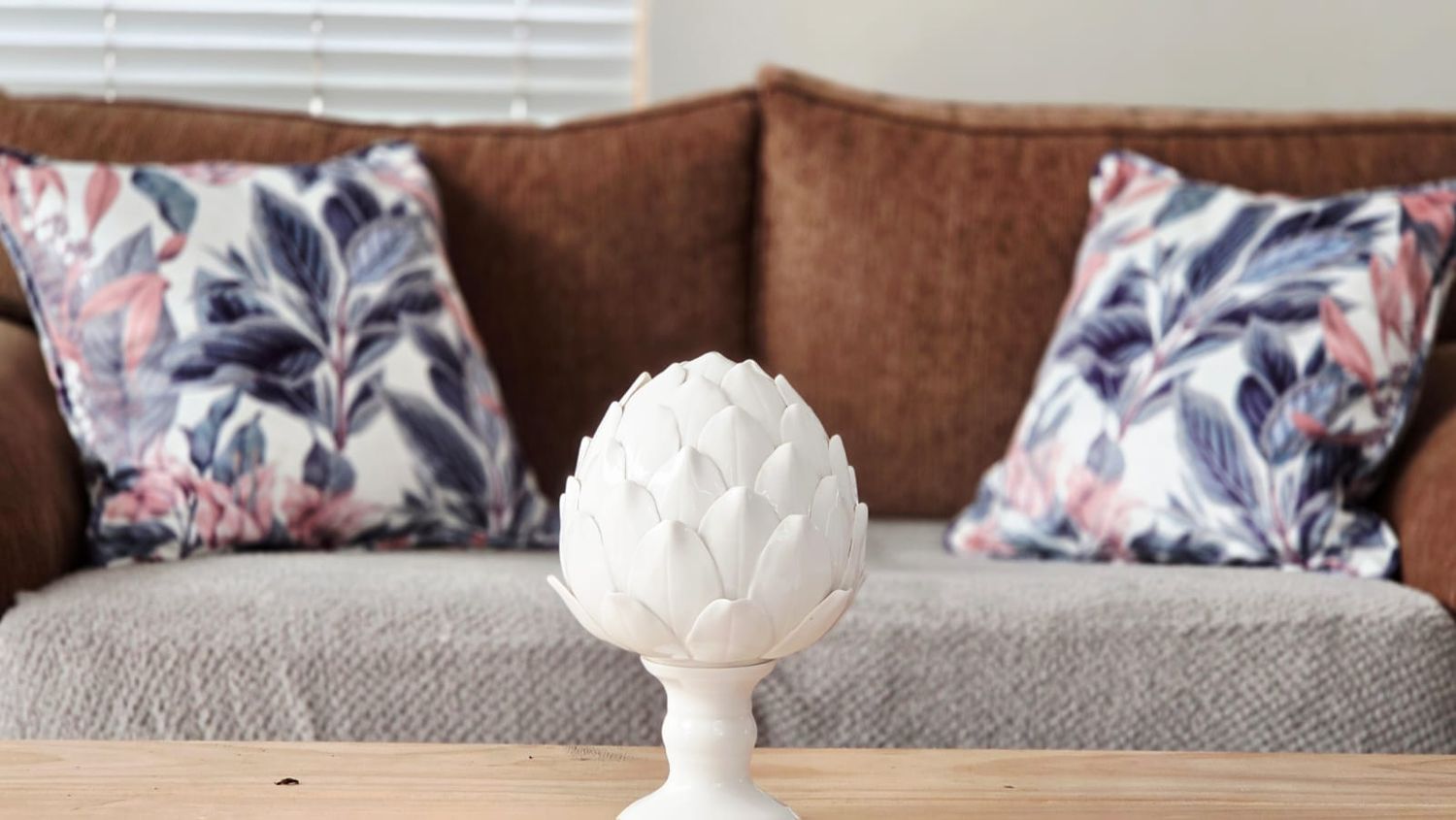 White ceramic artichoke figurine on wood table before brown sofa with floral pillows.