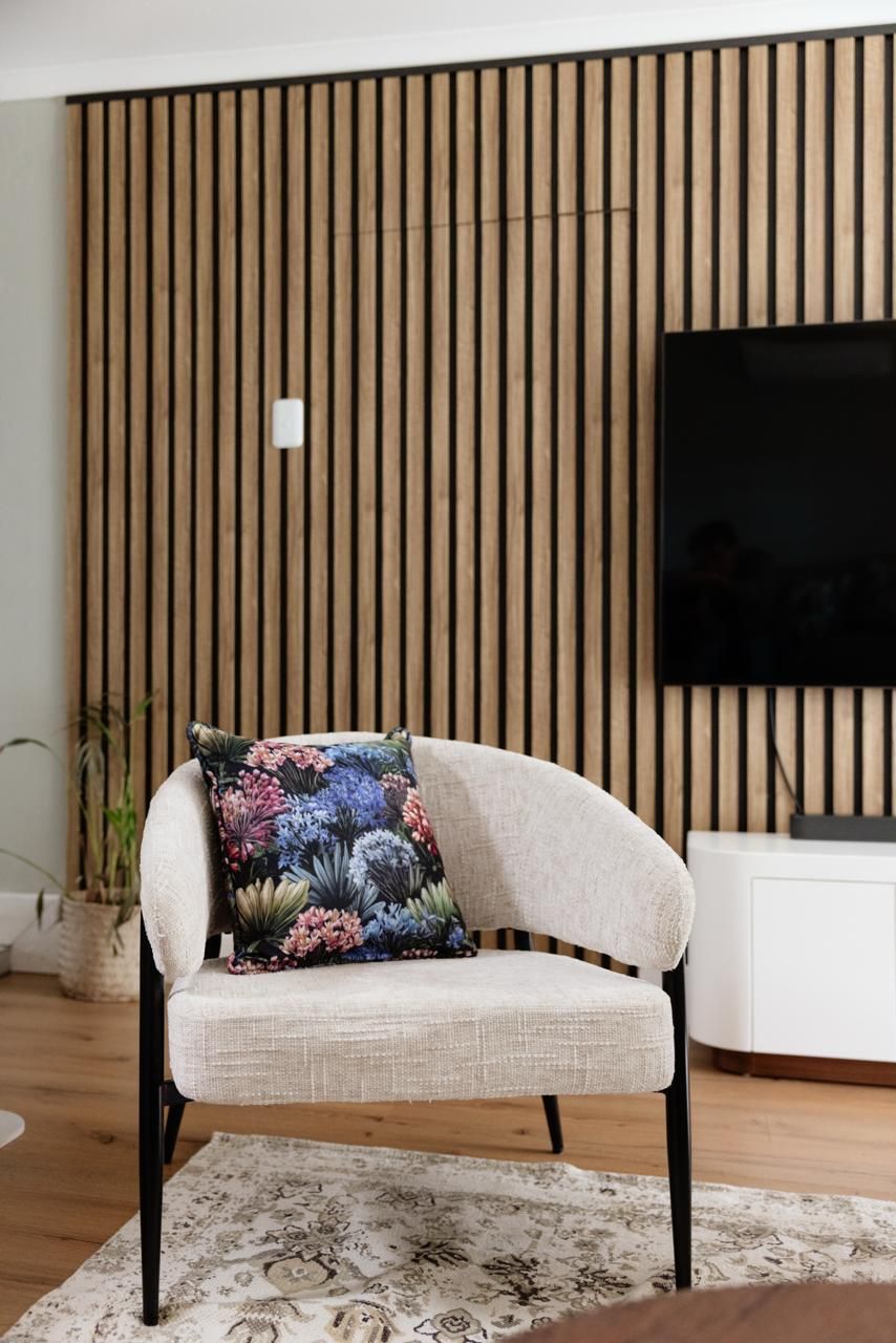 Modern living room featuring a beige accent chair with floral pillow, wooden slat wall, and wall-mounted TV on hardwood floors.
