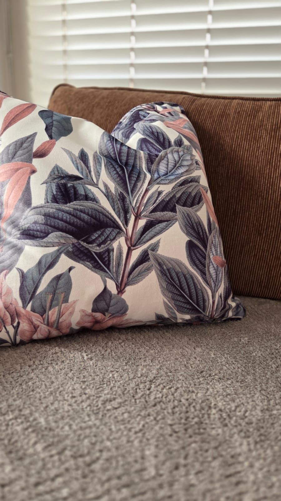 Decorative throw pillow with botanical leaf print in muted pink and purple tones resting on a brown corduroy sofa.